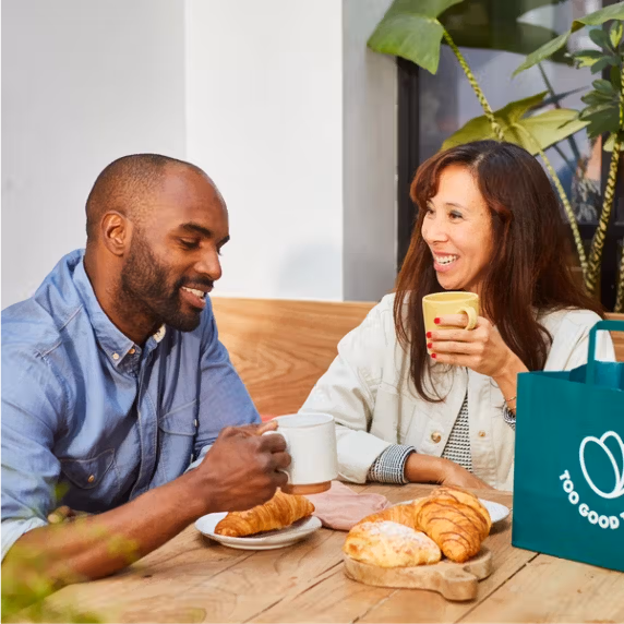 a man and woman sit at a table with a bag that says too good